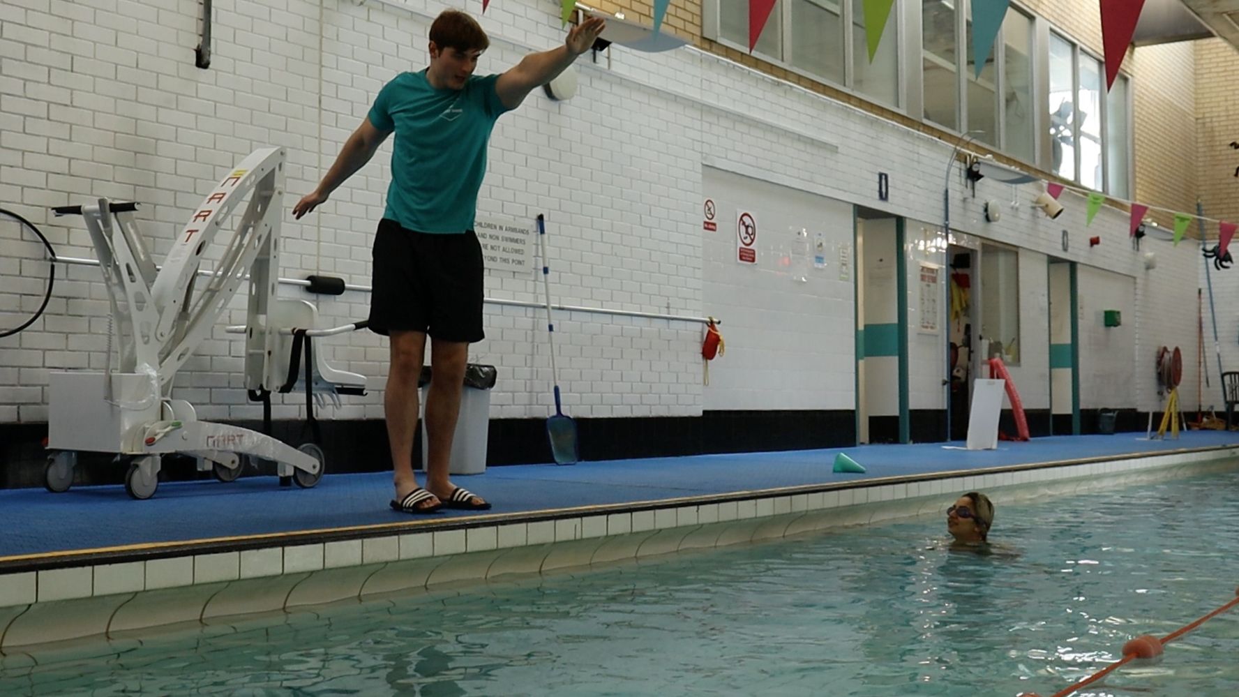 An instructor on the poolside demonstrating the arm pull while an adult learner watches from the water